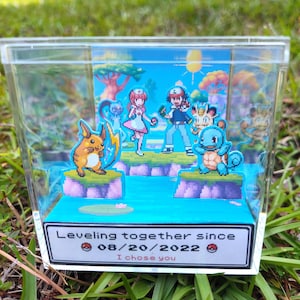 May include: A clear acrylic cube display box containing a multi-layered 3D diorama of pixelated characters including Raichu, Squirtle, Meowth, a nurse, and a trainer. The base features the text Leveling together since 08/20/2022 I chose you.
