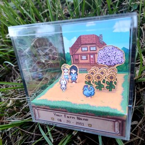 May include: A miniature diorama inside a clear acrylic box. The scene depicts a pixelated farm with a house, sunflowers, and characters. The base of the box has text that reads "Your Farm Name" and a date.
