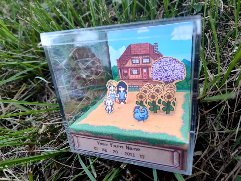 May include: A miniature diorama inside a clear acrylic box. The scene depicts a pixelated farm with a house, sunflowers, and characters. The base of the box has text that reads "Your Farm Name" and a date.