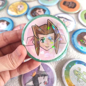 May include: A collection of colorful, round pinback buttons with various character illustrations. One button, held in a hand, features a character with spiky brown hair and green eyes. Other buttons display different characters and designs.