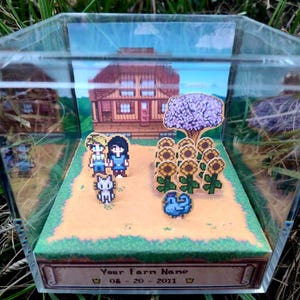 Könnte beinhalten: Miniatur-Diorama in einer transparenten Acrylbox. Die Szene zeigt einen pixeligen Bauernhof mit einem Haus, Sonnenblumen und Figuren. Die Basis der Box trägt den Text "Your Farm Name" und ein Datum. Das Diorama steht auf einem Bett aus grünem Gras.