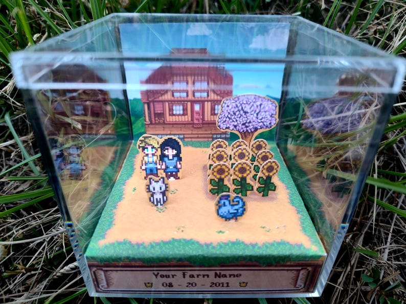May include: A miniature diorama inside a clear acrylic box. The scene depicts a pixelated farm with a house, sunflowers, and characters. The base of the box has text that reads "Your Farm Name" and a date. The diorama is set on a bed of green grass.