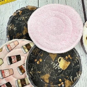 May include: A collection of round reusable coffee cup coasters. Some coasters are pink, others have a black background with gold coffee cup designs and the word "coffee". A measuring tape is in the background.