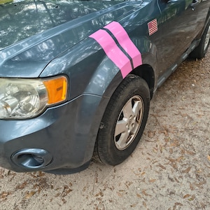 May include: A teal SUV with pink stripes on the front fender. The vehicle has a small American flag sticker and a silver wheel. The car is parked on a sandy surface.