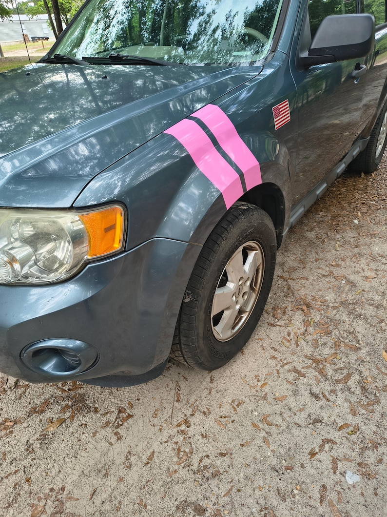 May include: A teal SUV with pink stripes on the front fender. The vehicle has a small American flag sticker and a silver wheel. The car is parked on a sandy surface.