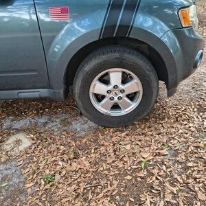 May include: A teal SUV with black racing stripes and an American flag sticker on the front wing. The vehicle has a silver wheel and a black tyre. The car is parked on a surface covered in fallen leaves.