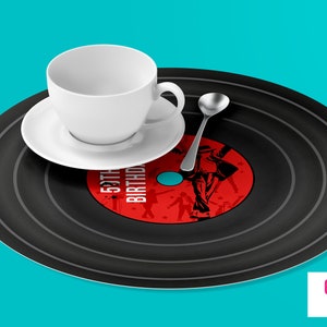 Puede incluir: Una bandeja de servir de vinilo negro en forma de disco con círculos concéntricos y un centro rojo con el texto "50th Birthday" y una silueta de una persona bailando. Una taza de té blanca y un platillo y una cucharadita de plata están en la bandeja.