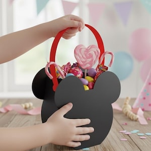 Puede incluir: Una cesta negra con forma de Mickey Mouse llena de caramelos y piruletas de colores. La cesta tiene un asa roja y la sostiene un niño. El fondo incluye decoraciones de fiesta y el texto "CRAFTY DIGITALS".