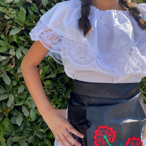 May include: A white off-the-shoulder blouse featuring a wide ruffled neckline with delicate white lace trim, paired with a black satin apron-style skirt adorned with embroidered red flowers and green stems, finished with a black lace border detail.