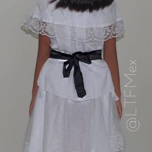 May include: A white dress featuring a ruffled capelet overlay with lace trim, a black ribbon tied at the waist, and a skirt with a lace-trimmed hem. The dress is shown from the back, and the text @LTFMex is visible on the right side of the image.