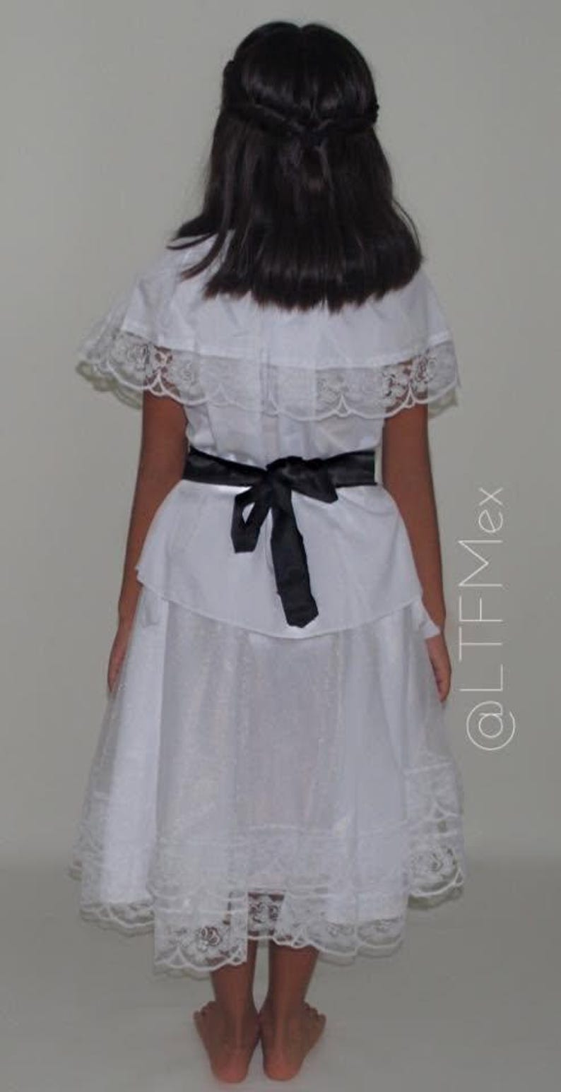 May include: A white dress featuring a ruffled capelet overlay with lace trim, a black ribbon tied at the waist, and a skirt with a lace-trimmed hem. The dress is shown from the back, and the text @LTFMex is visible on the right side of the image.