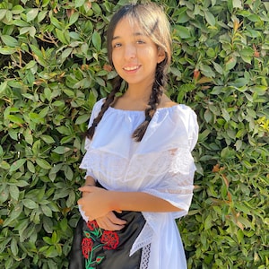 May include: A white off-the-shoulder dress featuring a ruffled neckline with white lace trim and short sleeves. A black apron with red and green floral embroidery and black lace edging is worn over the dress, which has a full-length white skirt.