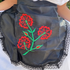 May include: A black apron featuring a central embroidered design of three red flowers with green stems and leaves. The apron is finished with a black lace trim along the bottom edge and is worn over a white garment with a white lace collar detail.