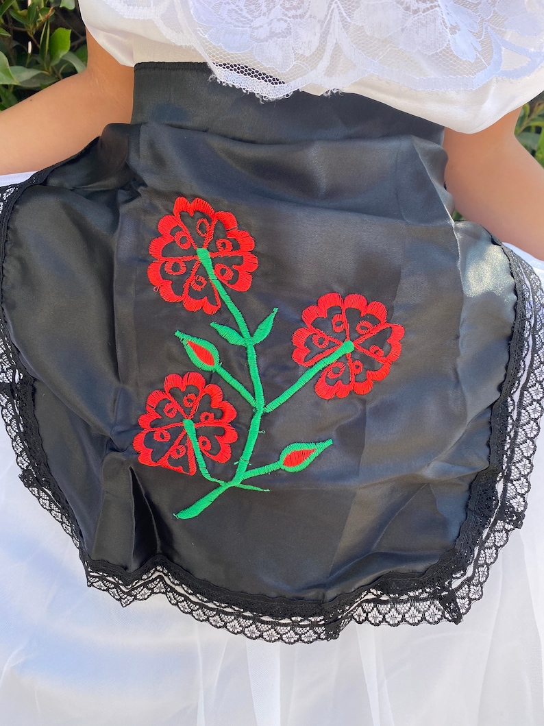 May include: A black apron featuring a central embroidered design of three red flowers with green stems and leaves. The apron is finished with a black lace trim along the bottom edge and is worn over a white garment with a white lace collar detail.