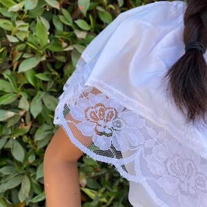 May include: A white blouse featuring a wide, off-the-shoulder ruffle collar adorned with a detailed floral lace trim along the edges. The fabric is smooth and lightweight, with the lace pattern showcasing intricate rose-like floral motifs.