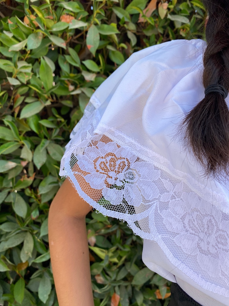 May include: A white blouse featuring a wide, off-the-shoulder ruffle collar adorned with a detailed floral lace trim along the edges. The fabric is smooth and lightweight, with the lace pattern showcasing intricate rose-like floral motifs.