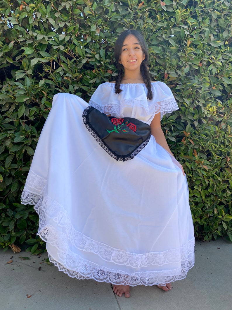 May include: A white off-the-shoulder dress featuring a ruffled neckline with lace trim, a long skirt with tiered lace detailing at the hem, and a black apron-style overlay adorned with an embroidered red rose design and black lace edging.
