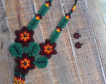 May include: A beaded necklace and matching earrings set. The necklace features large, dark red flowers with orange and yellow centers, green leaves, and a beaded fringe. The necklace and earrings are on a wooden surface.