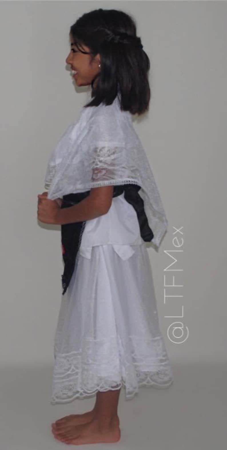 May include: A white dress featuring a sheer lace overlay with intricate patterns, a scalloped hem, and a black sash tied at the waist. The garment includes a matching white lace shawl draped over the shoulders. The text @LTFMex is visible.