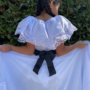 May include: A white off-the-shoulder dress featuring a wide ruffled neckline trimmed with white floral lace. The dress includes a black satin ribbon tied in a bow at the waist and a long, flowing skirt with a matching white lace border at the hem.