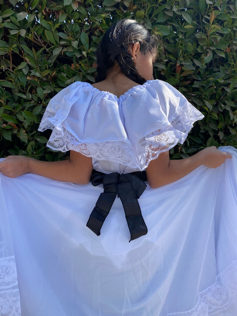 May include: A white off-the-shoulder dress featuring a wide ruffled neckline trimmed with white floral lace. The dress includes a black satin ribbon tied in a bow at the waist and a long, flowing skirt with a matching white lace border at the hem.