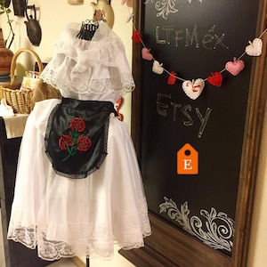 May include: A white dress featuring a lace-trimmed collar and hem, paired with a black apron decorated with an embroidered red flower design. The garment is displayed on a mannequin, showcasing intricate lace details and a contrasting black fabric.