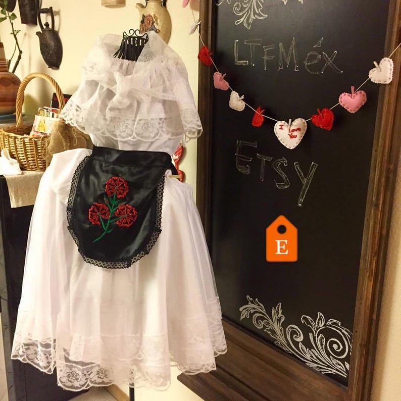 May include: A white dress featuring a lace-trimmed collar and hem, paired with a black apron decorated with an embroidered red flower design. The garment is displayed on a mannequin, showcasing intricate lace details and a contrasting black fabric.