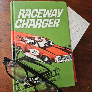May include: A green book with the title "Raceway Charger" in white letters. The cover features a red and black race car with the number 33 on the side. A pair of black glasses are lying on top of the book.