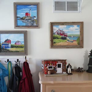 May include: Three framed paintings of lighthouses and coastal scenes hang on a white wall. A miniature lighthouse figurine and a small wooden model of a seaside shop are on a light brown dresser.
