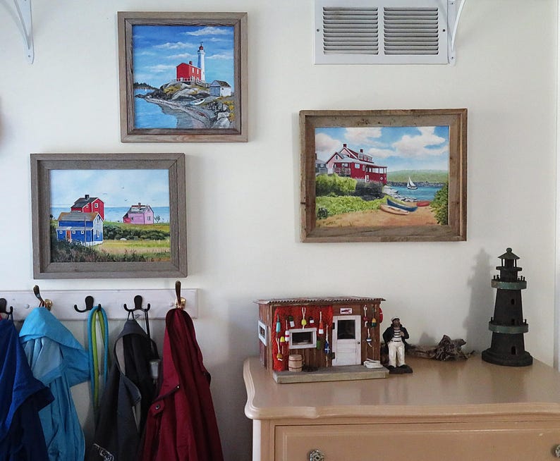 May include: Three framed paintings of lighthouses and coastal scenes hang on a white wall. A miniature lighthouse figurine and a small wooden model of a seaside shop are on a light brown dresser.