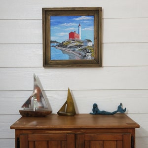 May include: A framed painting of a red lighthouse on a rocky shore with a blue sky and white clouds. The lighthouse has a white tower and a red roof. There are two small boats in the water.