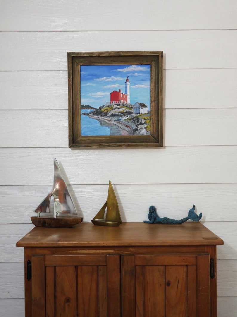 May include: A framed painting of a red lighthouse on a rocky shore with a blue sky and white clouds. The lighthouse has a white tower and a red roof. There are two small boats in the water.