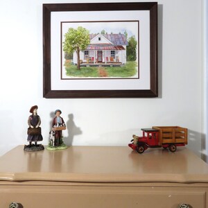 May include: A framed watercolor painting of a white farmhouse with a red roof and a porch. The house is surrounded by green trees and grass. The painting is on a white wall with a brown wooden frame.