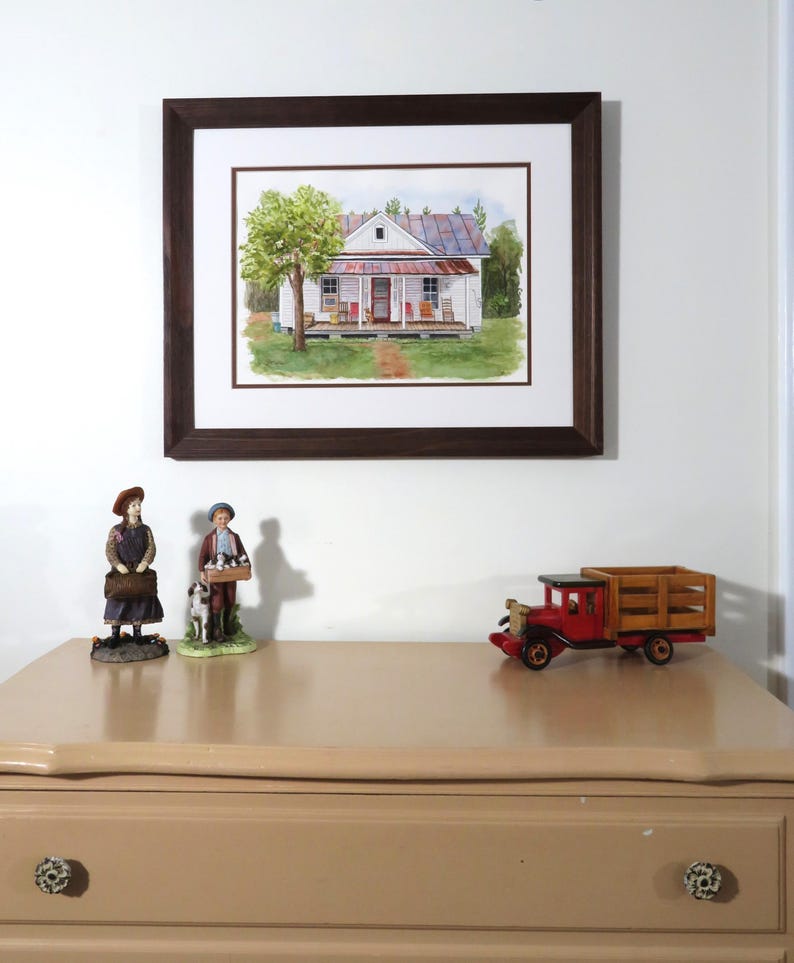 May include: A framed watercolor painting of a white farmhouse with a red roof and a porch. The house is surrounded by green trees and grass. The painting is on a white wall with a brown wooden frame.