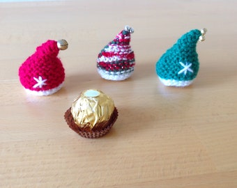 May include: Three miniature crocheted elf hats in red, green, and a red/white/green striped pattern, each with a white star or trim. A wrapped chocolate candy sits in front of the hats on a wooden surface. The hats have small gold bells.