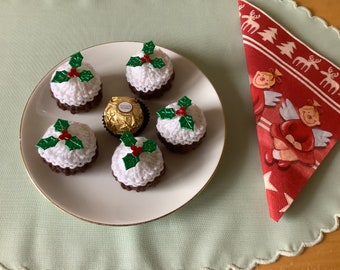 May include: Five white crocheted Christmas pudding ornaments with green holly and red berries on a white plate. A single chocolate candy is in the center of the plate.