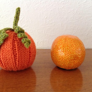 Könnte beinhalten: Ein gestrickter orangefarbener Kürbis mit grünen Blättern und eine echte Orange daneben.