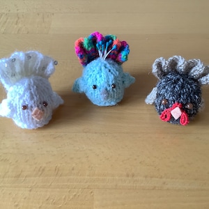 May include: Three knitted bird ornaments. The first bird is white with a white and silver crown. The second bird is light blue with a colourful crown. The third bird is grey with a brown beak and a grey crown.