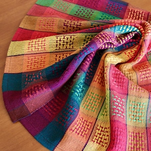 May include: A colorful, woven textile with a vibrant array of hues. The fabric features a checkered pattern with squares of red, orange, yellow, green, blue, and purple. The weave creates a textured appearance, with a soft, draped form.