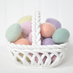 May include: A white ceramic basket filled with pastel coloured felt Easter eggs. The eggs are in shades of pink, yellow, green, and purple.