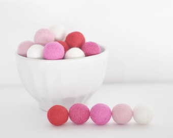 Könnte beinhalten: Eine weiße Schüssel gefüllt mit rosa, roten und weißen Filzkugeln. Eine Reihe von Filzkugeln in den gleichen Farben sind auf einer weißen Oberfläche aufgereiht.