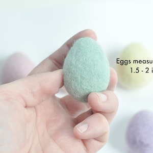 May include: A hand holding a light green felt egg.  The egg measures approximately 1.5-2 inches long.  There are other felt eggs in the background, one yellow and one purple.
