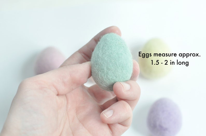 May include: A hand holding a light green felt egg.  The egg measures approximately 1.5-2 inches long.  There are other felt eggs in the background, one yellow and one purple.