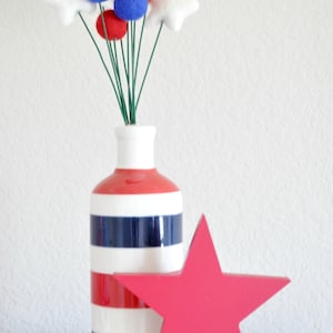 K&ouml;nnte beinhalten: Eine rot-wei&szlig;-blau gestreifte Keramikvase mit einem Strau&szlig; roter, wei&szlig;er und blauer Filzblumen. Ein gro&szlig;er roter Stern steht vor der Vase.