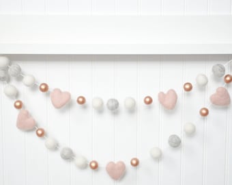 Op de afbeelding: Een guirlande met witte en grijze viltballen en roze vilt harten, afgewisseld met roosgoudkleurige kralen. De guirlande hangt aan een wit rek.