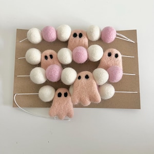 May include: A garland of white and pink felt balls with pink felt ghost ornaments. The garland is strung on white thread and is displayed on a brown cardboard background.