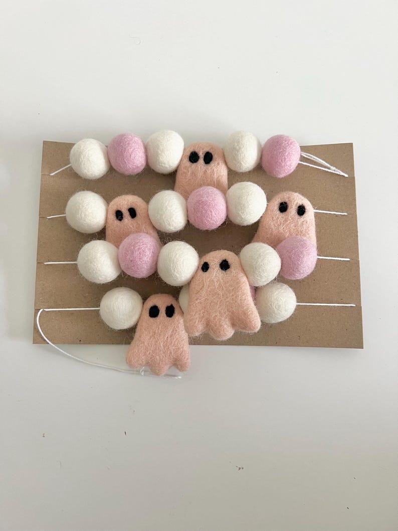 May include: A garland of white and pink felt balls with pink felt ghost ornaments. The garland is strung on white thread and is displayed on a brown cardboard background.