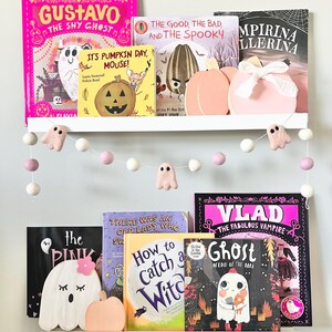 May include: A white shelf with a collection of children's books featuring Halloween themes. The books include titles like "Gustavo the Shy Ghost", "It's Pumpkin Day, Mouse!" and "Vlad the Fabulous Vampire". There are also several small, white, felt ghost decorations hanging from the shelf.