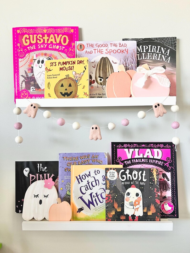 May include: A white shelf with a collection of children's books featuring Halloween themes. The books include titles like "Gustavo the Shy Ghost", "It's Pumpkin Day, Mouse!" and "Vlad the Fabulous Vampire". There are also several small, white, felt ghost decorations hanging from the shelf.