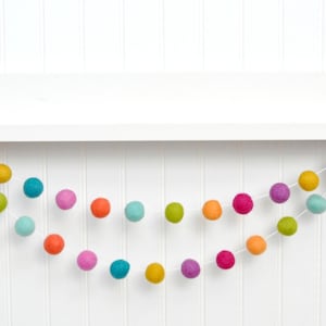 K&ouml;nnte beinhalten: Eine bunte Filzkugelgirlande mit verschiedenen Farben, darunter Rosa, Orange, Gelb, Gr&uuml;n, Blau und Lila. Die Girlande ist auf einer wei&szlig;en Schnur aufgef&auml;delt und h&auml;ngt an einem wei&szlig;en Regal.
